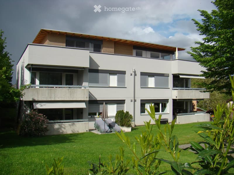 Two-story building, grey color, wooden roof, several balconies, lawn with plants and flowers, trees
