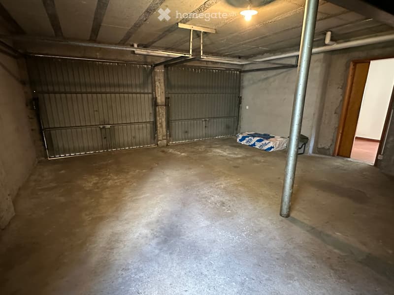 Unfurnished Room. Empty garage with concrete floor, metal doors, and a single support column. A folded mattress and a small trash bin are visible in the corner.