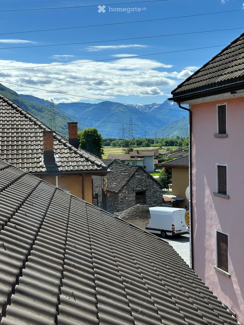 Building. Roof tops of houses in a village with a mountainous backdrop, featuring tiled roofs, chimneys, and a white van parked on a street.