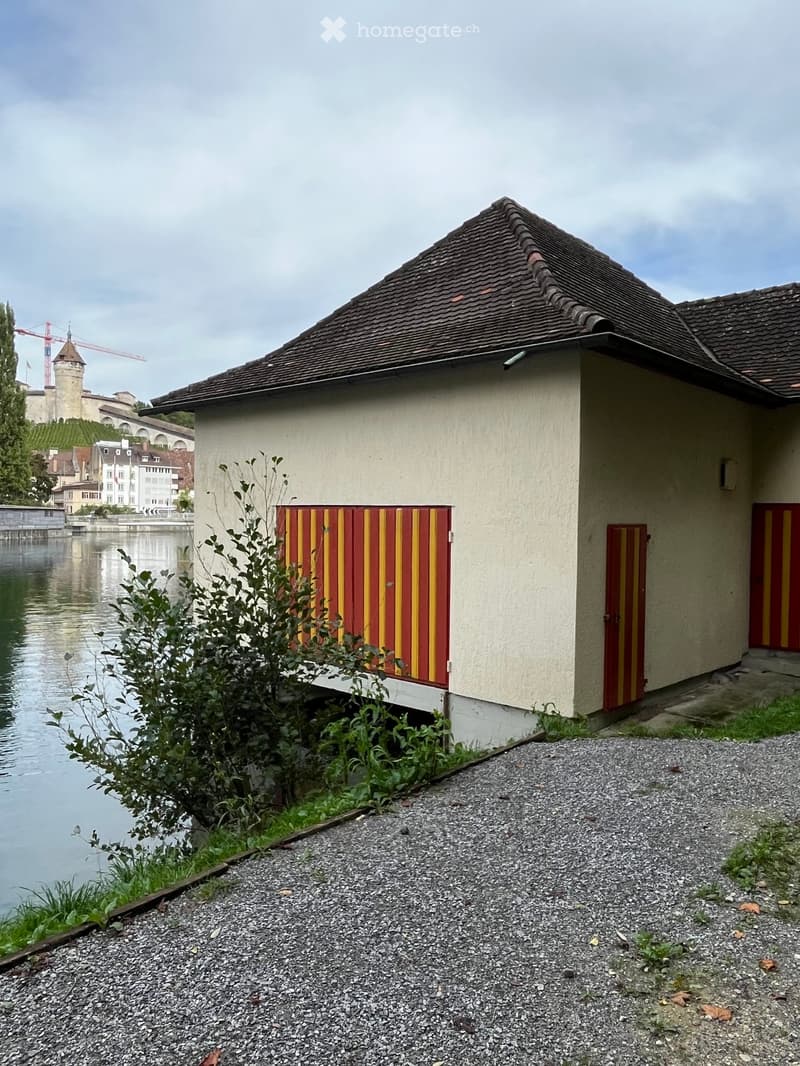 Building. A small, single-story building with a dark tiled roof and light-colored walls, featuring red and yellow striped doors and shutters. Located near a body of water with a view of a distant castle and town.