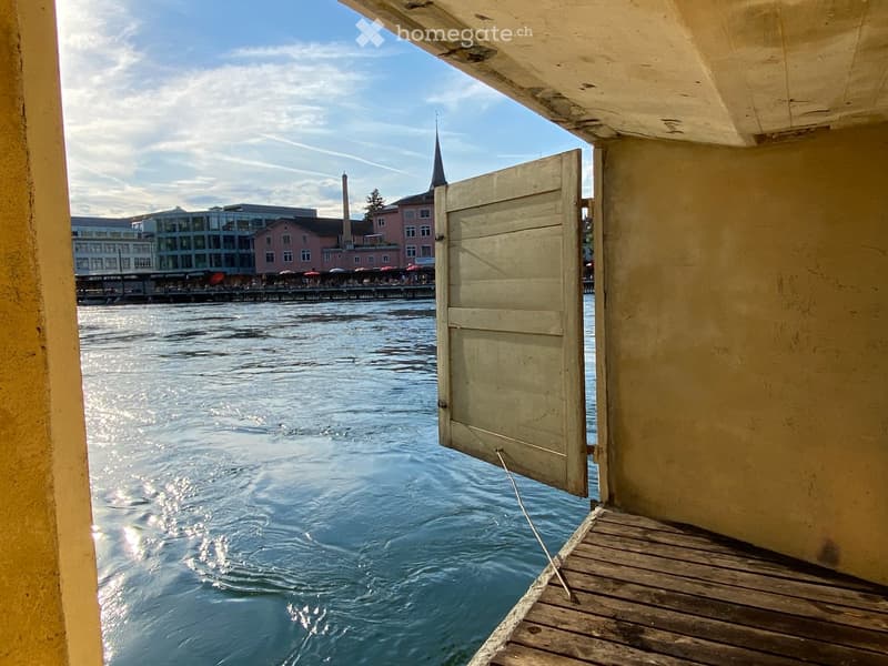 Balcony / Terrace. Open wooden balcony overlooking a body of water with a cityscape in the background, featuring a closed wooden door and a wooden deck.
