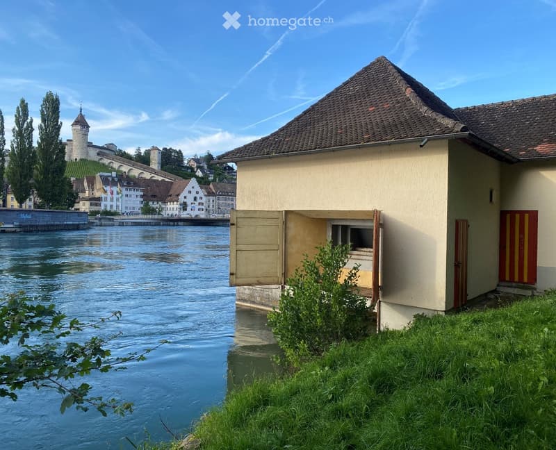 Building. A small beige building with a tiled roof sits near a river, with a view of a town and a castle in the background.