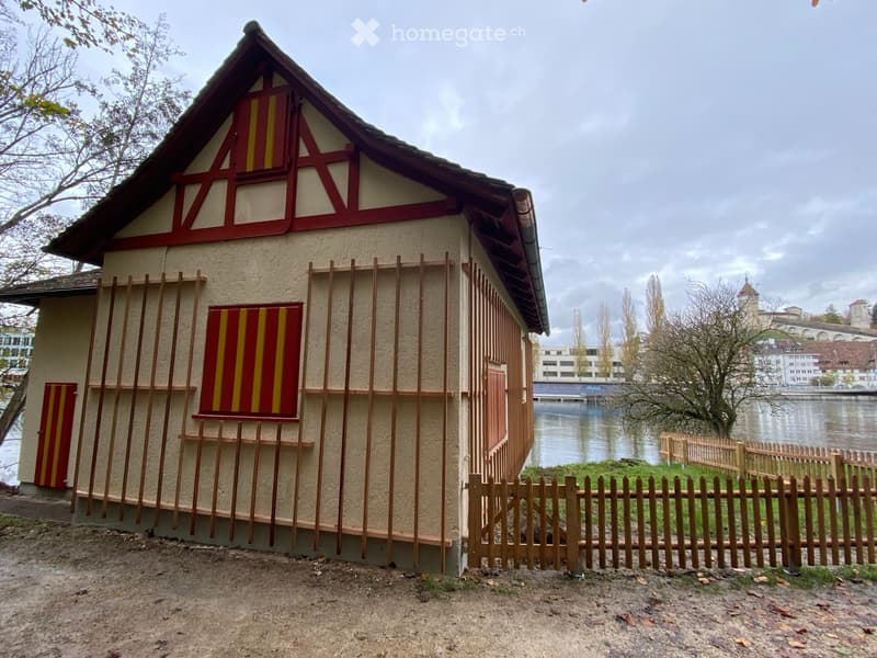 Building. A small, traditional wooden house with a pitched roof and decorative red and yellow panels, situated near a river with a wooden fence in front.