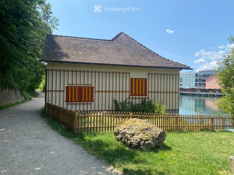 House. A small, single-story house with a tiled roof and wooden fence, situated near a waterfront with a view of the water and distant buildings.