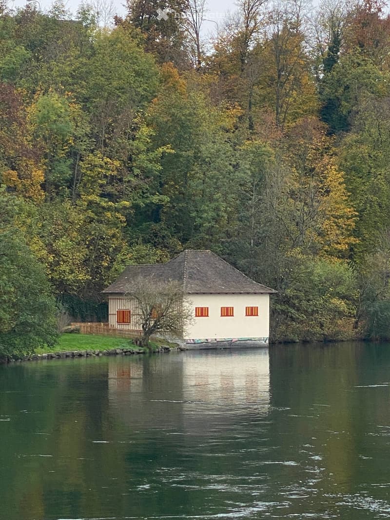 Building. A small, white wooden house with a sloped roof and three windows, situated on the bank of a calm river surrounded by trees.