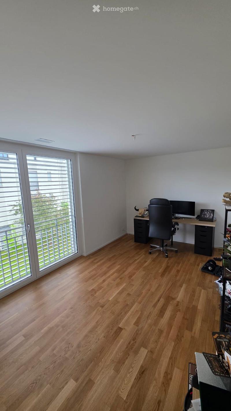 Furnished Room. A modern, well-lit room with wooden flooring, a large window with blinds, and a desk setup including a chair, computer, and shelves.