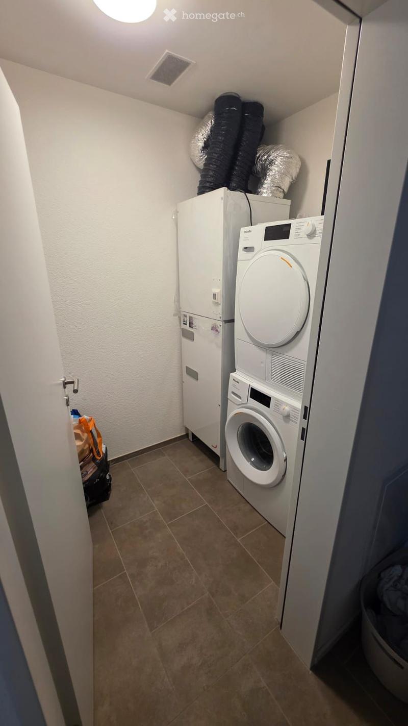 Furnished Room. A small, well-lit laundry room with a refrigerator, washing machine, and dryer. The floor is tiled, and the walls are white.