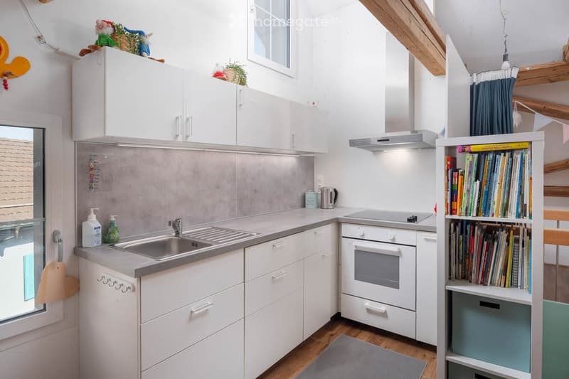 Kitchen. Modern kitchen with white cabinets, stainless steel sink, oven, and a bookshelf filled with books. The space features a wooden floor and a small window above the sink.