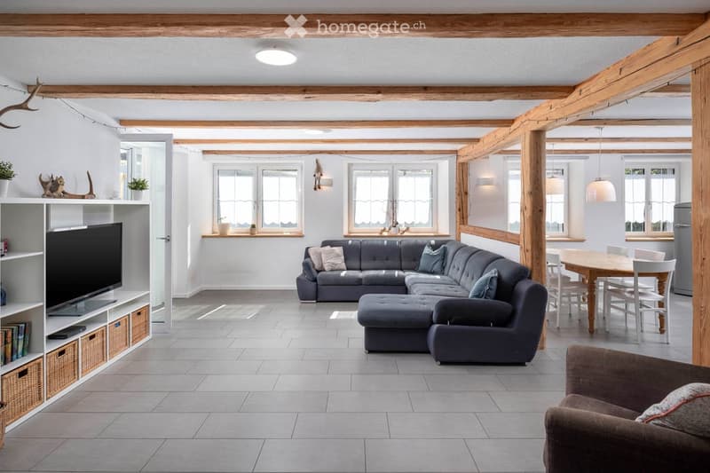 Furnished Room. Open-concept living and dining area with exposed wooden beams, grey sectional sofa, ottoman, dining table with chairs, and a TV on a shelf.