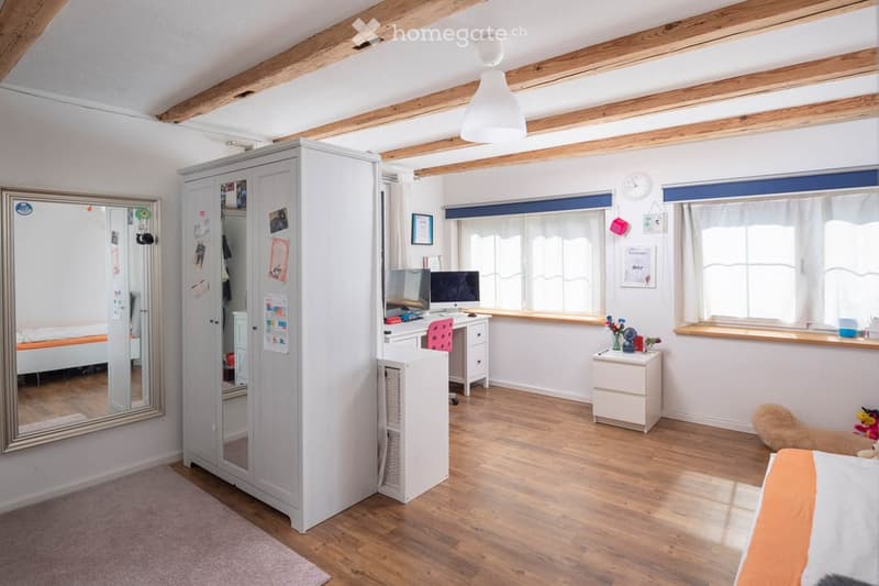 Furnished Room. A well-lit bedroom with wooden floors, exposed wooden beams, a large wardrobe, a desk with a computer, a nightstand, and a bed with orange bedding.