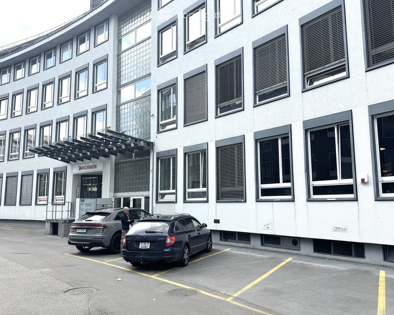 Building. Modern multi-story building with large windows and a covered entrance, with two parked cars in front.