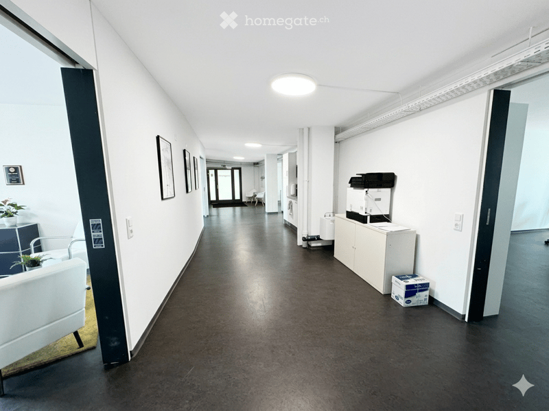 Corridor / Hallway. Modern hallway with white walls, dark polished floor, and minimal furnishings including a small seating area and a cabinet with office equipment.