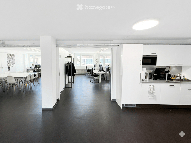 Furnished Room. Modern office space with a kitchen area, white cabinets, microwave, coffee maker, sink, and a dining area with a table and chairs. There are also workstations with laptops and screens.