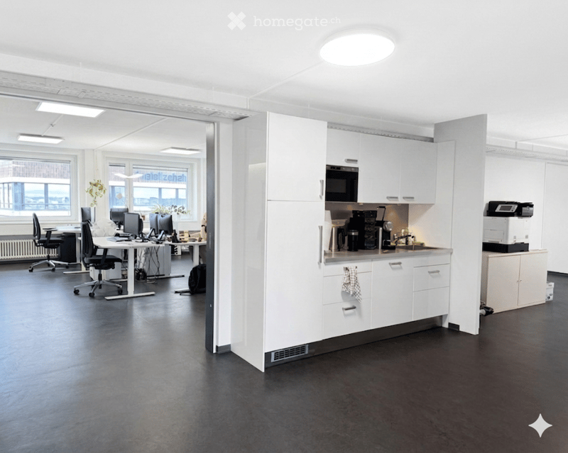 Furnished Room. Modern office space with a kitchen area, featuring white cabinets, a microwave, a printer, and a workspace with chairs and tables.