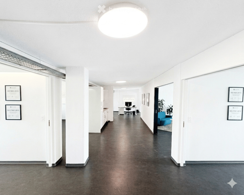 Corridor / Hallway. Modern hallway with white walls, dark polished floor, and a large central light fixture. Visible ISO certification plaques on walls, with a glimpse of an office area featuring a desk, chair, and blue couch in the background.