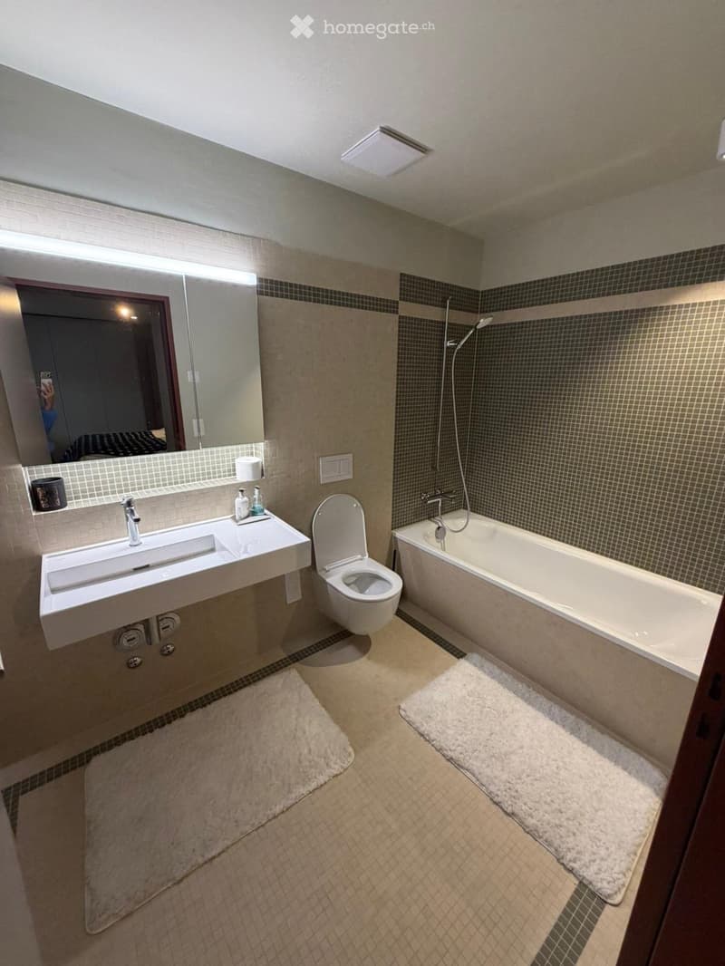 Bathroom. Modern bathroom with a white sink, toilet, bathtub, and large mirror. Beige tiles and two white rugs on the floor.