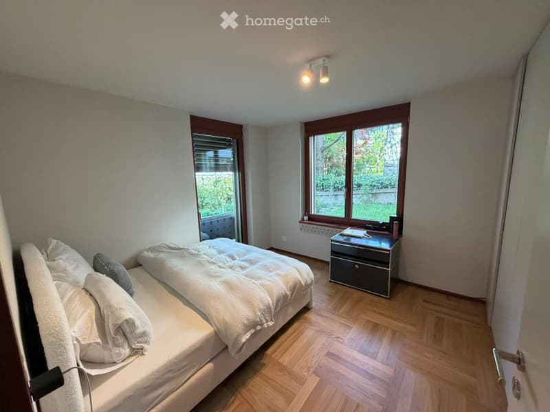 Bedroom. Cozy bedroom with wooden flooring, large windows, a neatly made bed, and a nightstand with a lamp.