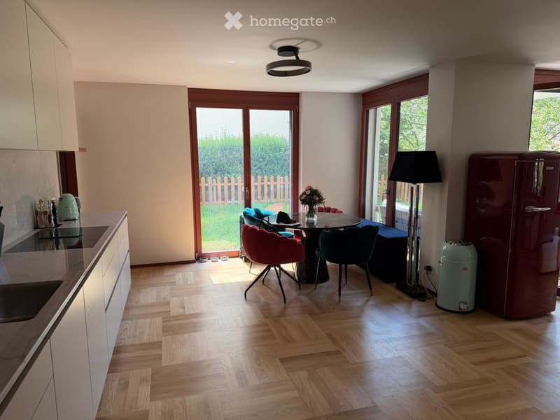 Furnished Room. Open-plan kitchen and dining area with wooden flooring, modern kitchen cabinets, a round dining table with chairs, a floor lamp, and a refrigerator.