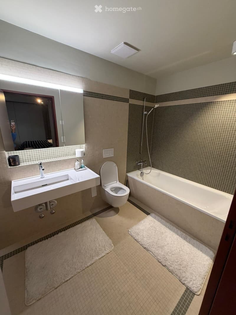 Bathroom. Modern bathroom with a white sink, toilet, bathtub, and large mirror. Beige tiles and white rugs are present.