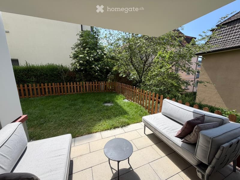 Garden / Outdoor Area. Outdoor patio with a grey sofa, small round table, and chair, surrounded by a wooden fence and green lawn.