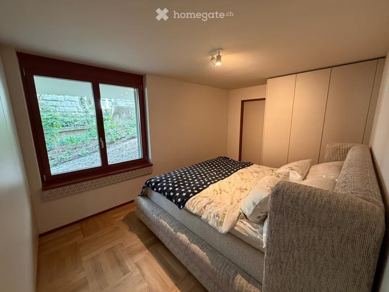 Bedroom. Cozy bedroom with a grey upholstered bed, patterned blanket, pillows, and a built-in wardrobe. Large window offers a view of a garden.
