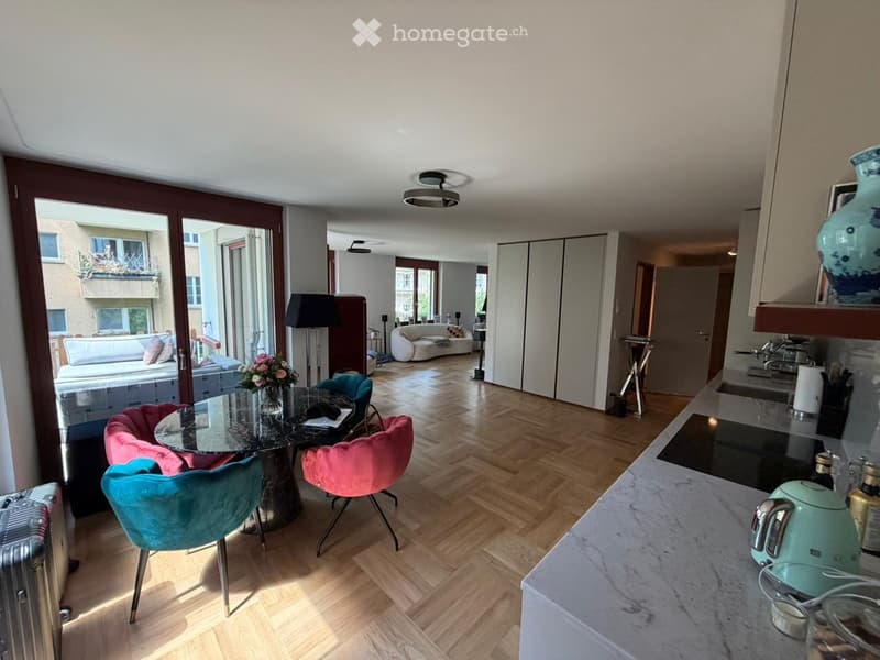 Furnished Room. Modern, well-lit living and dining area with wooden flooring, a glass dining table with colorful chairs, a sofa, and a kitchen counter with appliances.