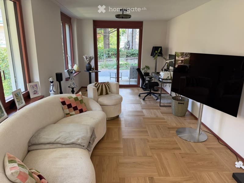 Living Room. Modern living room with wooden flooring, large windows, a curved sofa, armchair, coffee table, TV screen, and decorative items.