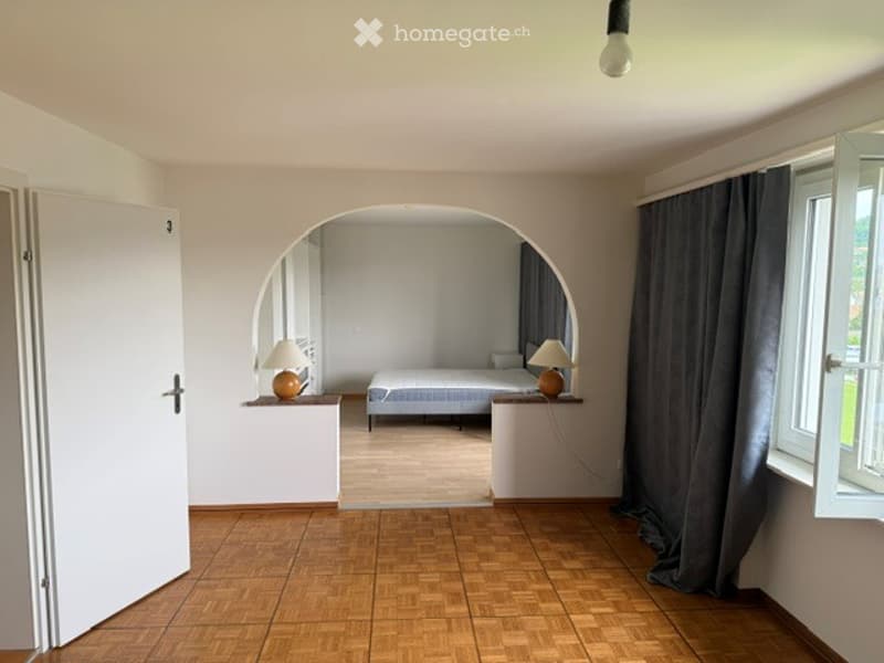 Furnished Room. A furnished room with a bed, nightstands, and lamps. The room features wooden flooring and large windows with curtains.