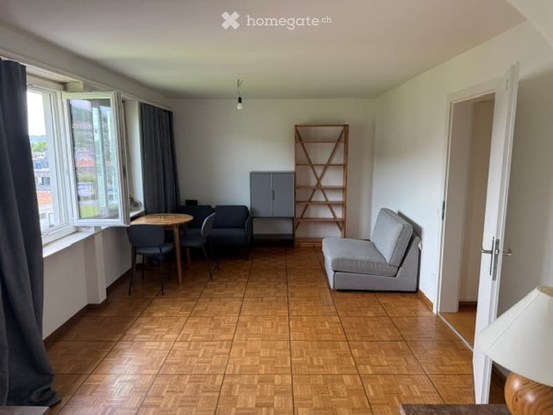 Furnished Room. Bright living room with wooden flooring, large windows with curtains, a grey sofa, a small table with chairs, a wooden shelf, and a built-in cabinet.