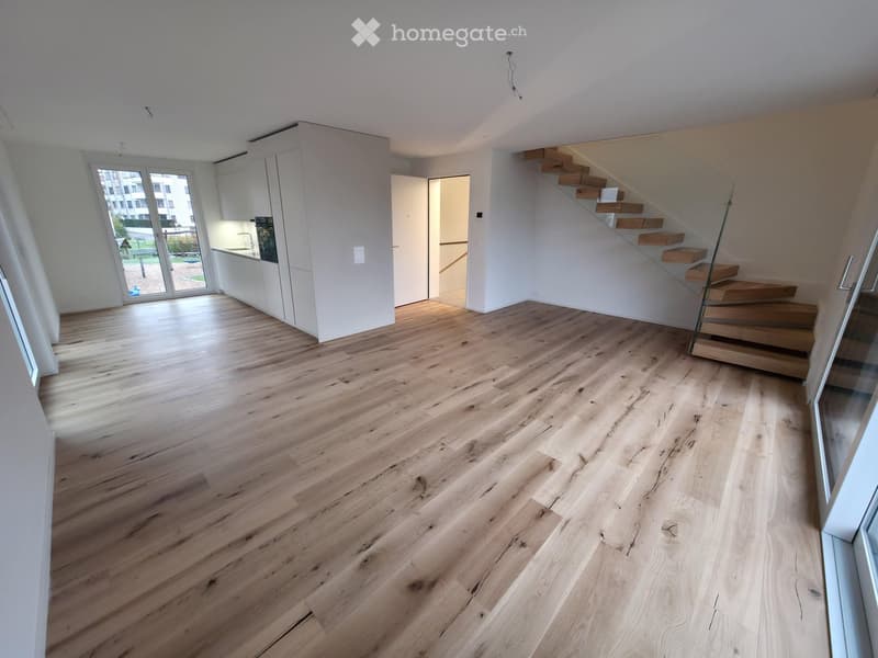 Unfurnished Room. Open-plan space with light wooden flooring, white walls, and a staircase leading upwards. The area includes a kitchen area with white cabinets and a doorway leading to a balcony or outdoor space.