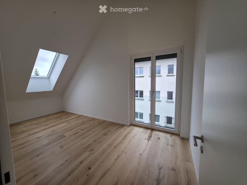 Unfurnished Room. Empty attic room with wooden flooring, white walls, large skylight window, and sliding glass door leading to a view of neighboring buildings.