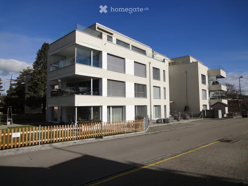 Building. Modern two-story residential building with white facade, large windows, and balconies. Features a wooden fence and small garden area in front.