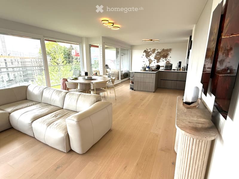 Furnished Room. Modern living room with a white leather sofa, coffee table, kitchen island, dining area, and large windows offering a view of greenery and construction.