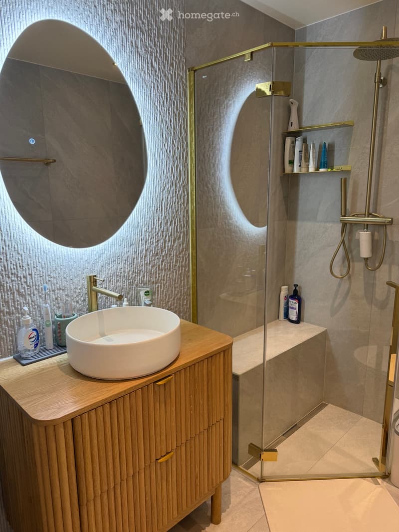 Bathroom. Modern bathroom with a wooden vanity, white sink, round mirror, glass-enclosed shower with gold fixtures, and various toiletries.