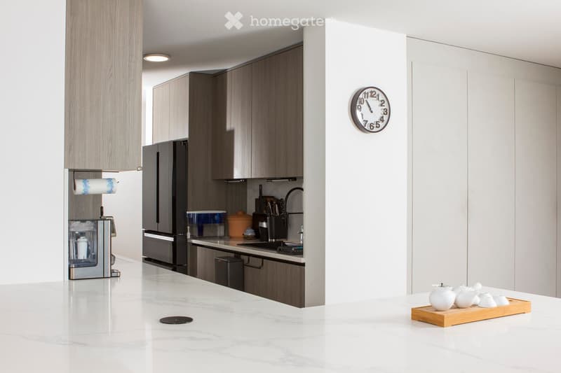 Kitchen. Modern kitchen with white marble countertops, dark wood cabinets, stainless steel appliances, and a coffee maker on the counter. A clock is mounted on the wall, and a tray with teapots and cups is placed on the island.