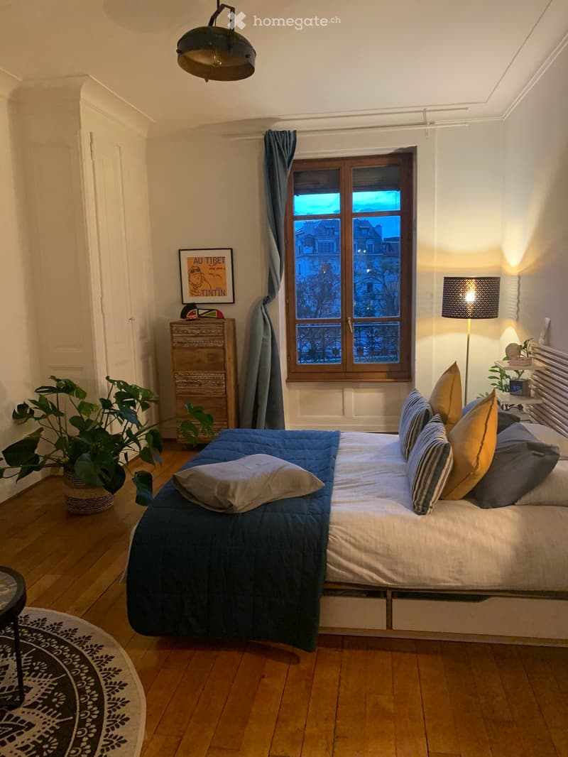 Bedroom. Cozy bedroom with wooden floor, large window with view, bed with blue and white bedding, nightstand with lamp, plant, and decorative items.