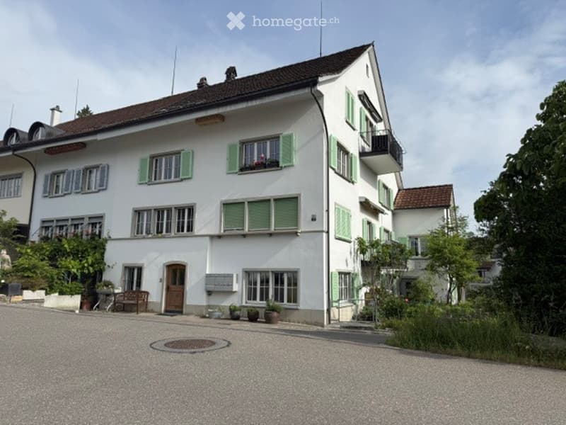 House. A two-story white house with green shutters and a brown roof, featuring multiple windows and a small balcony on the upper floor. The entrance has a wooden door and a small bench nearby.