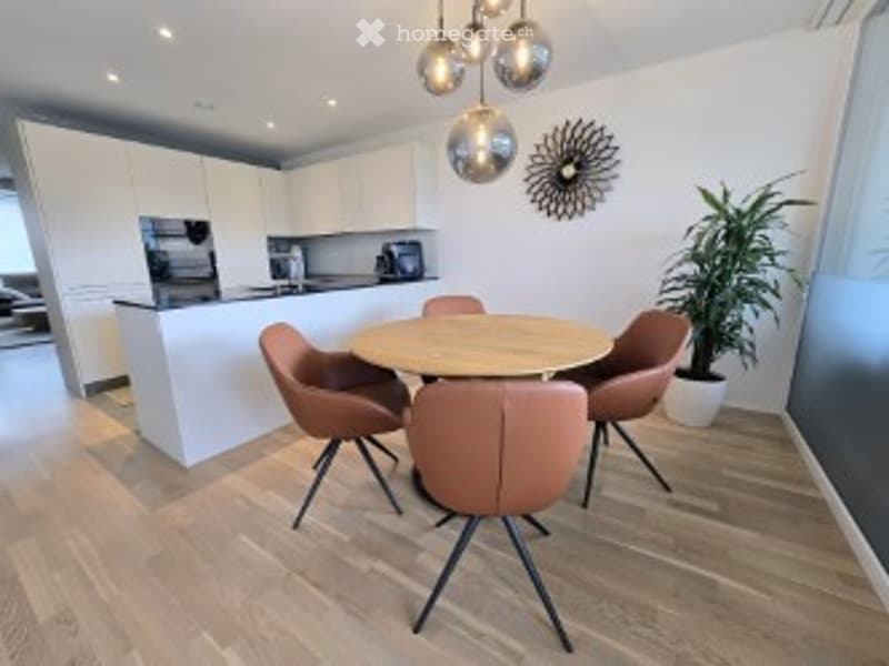 Furnished Room. Modern dining area with a round wooden table, four brown chairs, white kitchen island, pendant lights, and a potted plant.