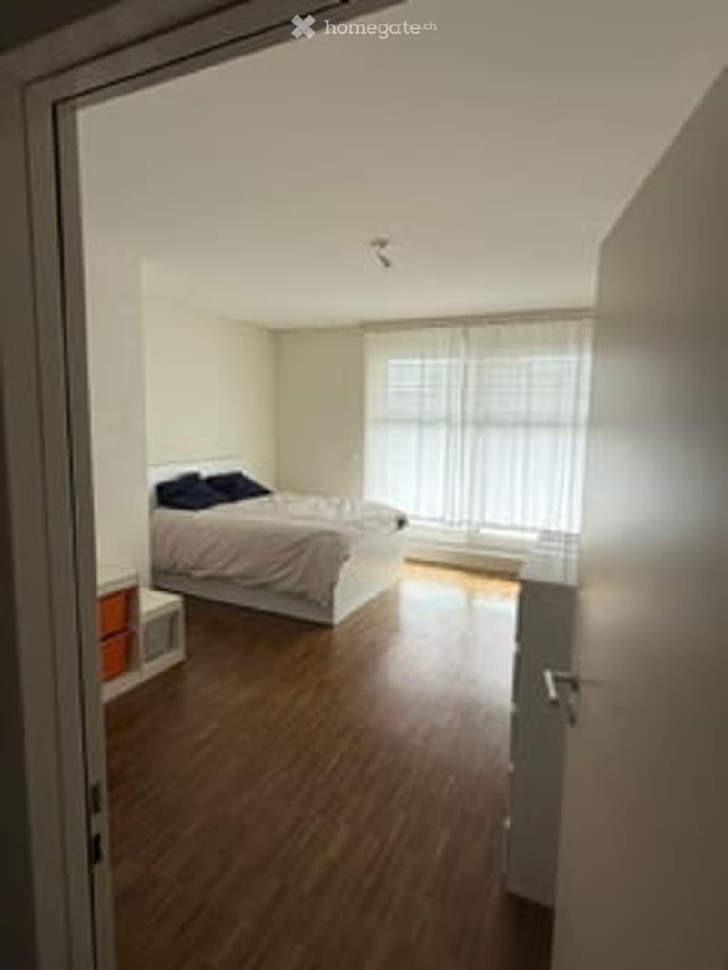 Bedroom. Bright bedroom with wooden flooring, large window with white curtains, bed with dark blue pillows, nightstand, and lamp.