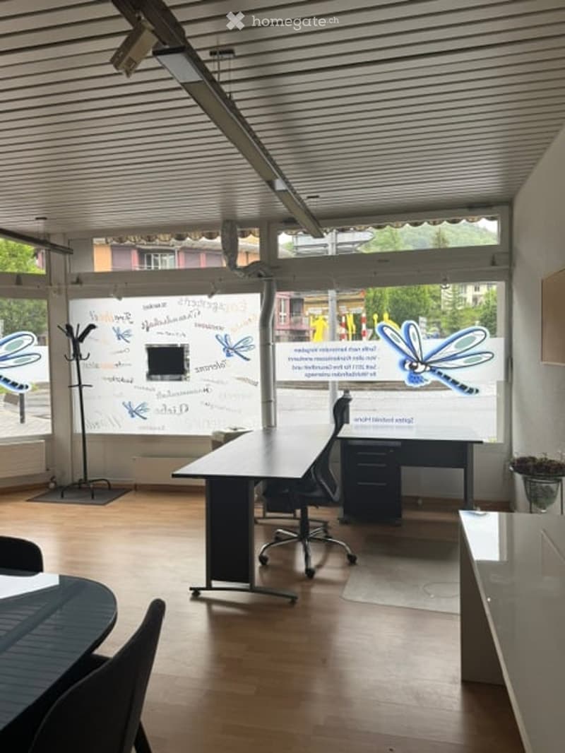 Furnished Room. Bright, modern office space with large windows, wooden flooring, a desk, chairs, a coat rack, and a screen displaying artwork.