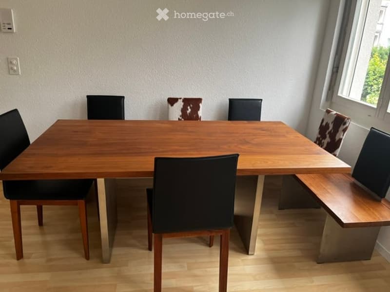 Dining area with a wooden table and multiple chairs, including a bench seat, set against a light-colored wall with a window to the right.