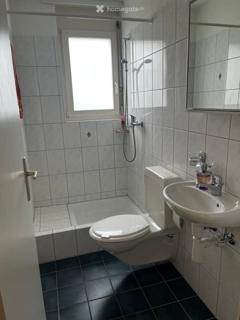 Bathroom with white tiled walls, black tiled floor, toilet, sink, shower, and a window above the shower area.