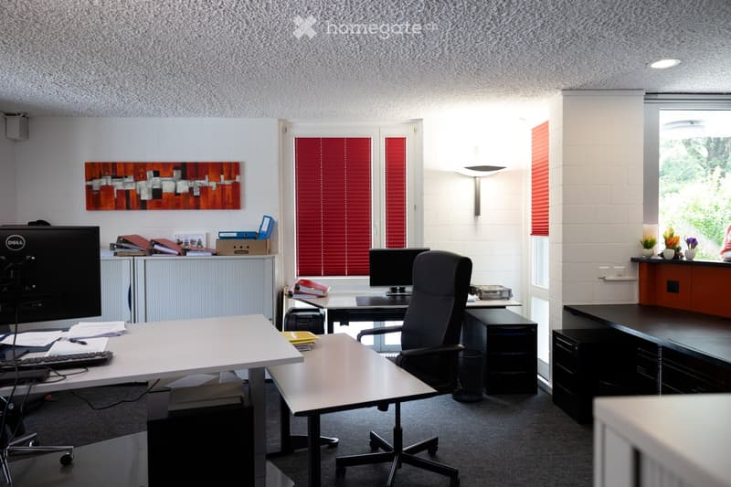 Furnished Room. Office space with desks, chairs, computers, and a printer, featuring a modern aesthetic with white brick walls and large windows with red blinds.