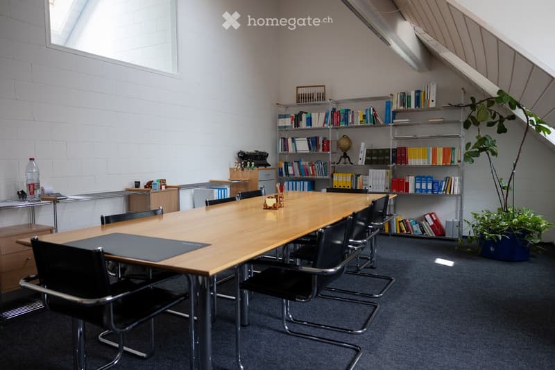 Furnished Room. A modern office or meeting room with a long wooden table surrounded by black chairs, metal shelving units filled with books and binders, a potted plant, and a globe on a stand. The room features exposed brick walls and large windows with b