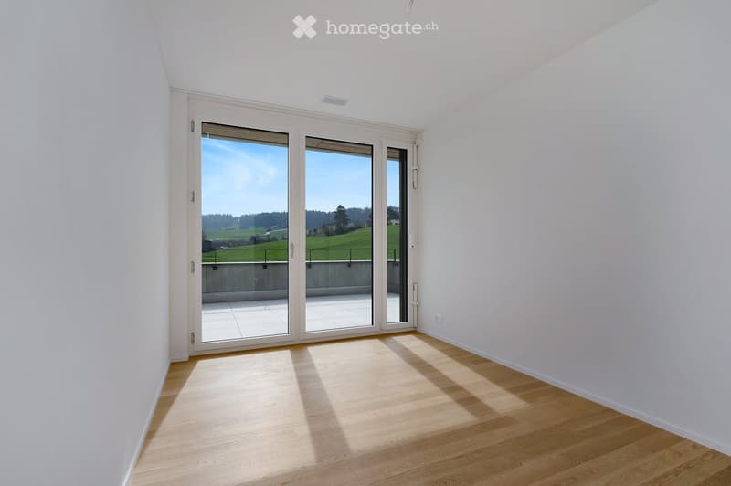 Unfurnished Room. Bright, empty room with white walls, wooden flooring, and large sliding glass doors leading to a balcony with a scenic view of green fields and trees.