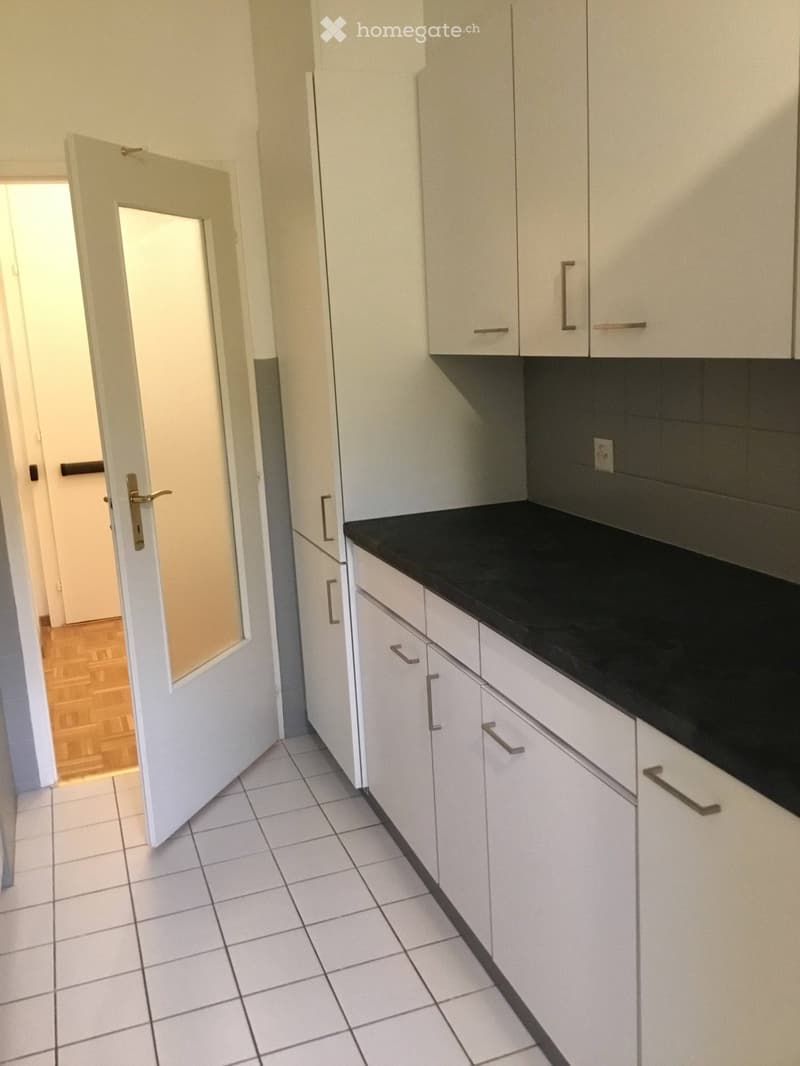 Unfurnished Room. Empty kitchen with white cabinets, black countertop, and tiled floor. An open door leads to another room with wooden flooring.