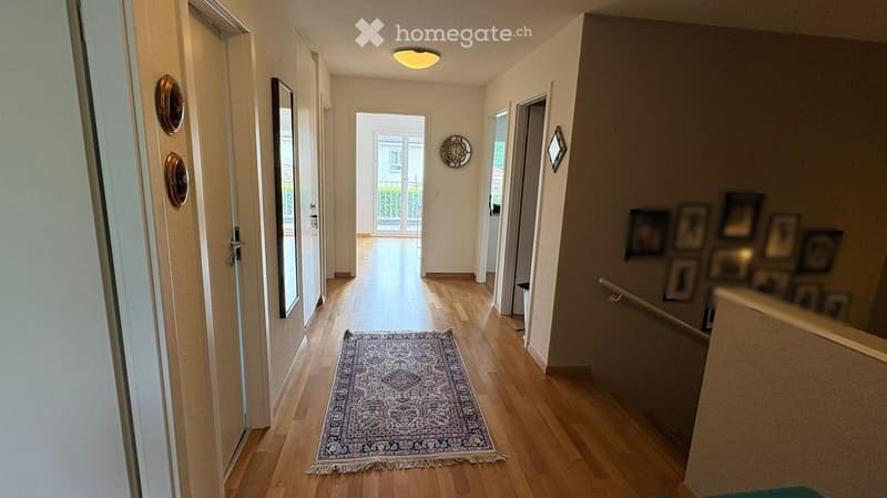 Corridor / Hallway. Bright hallway with wooden flooring, a patterned rug, and several doors leading to different rooms. A staircase is visible on the right side with framed pictures on the wall.