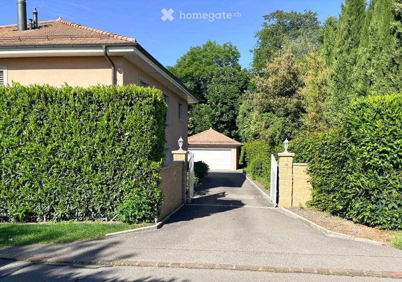House. A two-story house with a tiled roof, beige exterior, and a well-maintained hedge-lined driveway leading to a garage.