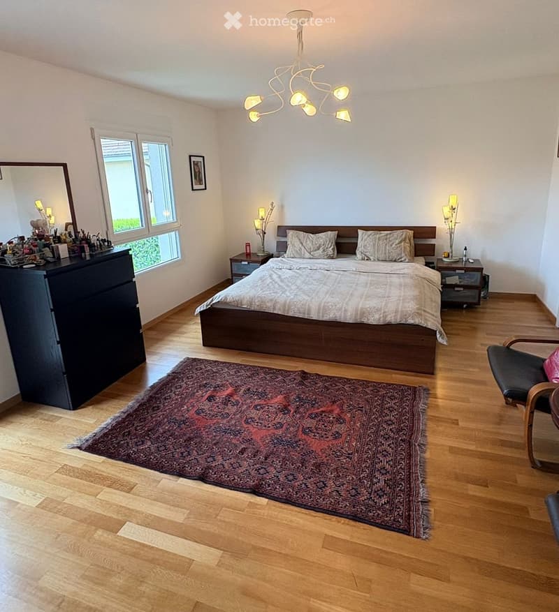 Bedroom. Spacious bedroom with wooden flooring, large bed with pillows and bedding, nightstands with lamps, a patterned rug, a black dresser, and a modern chandelier.