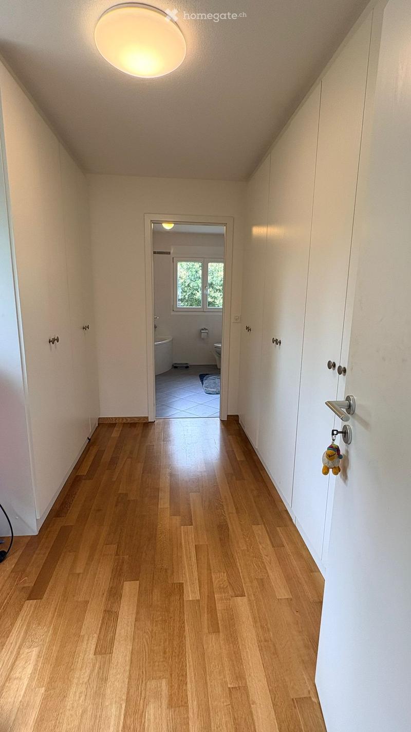 Corridor / Hallway. Bright hallway with wooden flooring, white walls, and a round ceiling light. A small bathroom is visible at the end of the hallway.